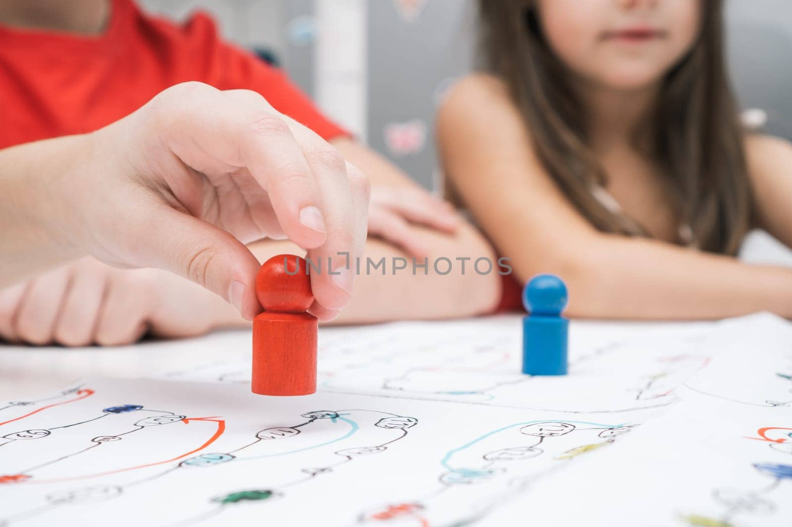 Little friends play board game on paper map with dice and chips at table. Hand of player hold red figure closeup, selective focus. Home leisure, playtime. Entertaining interesting table game.
