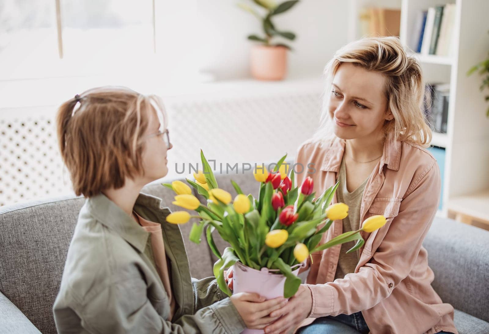 Mother and daughter sitting on the sofa with tulips in their hands indoor. Concept of the mother's day, birthday and motherhood.