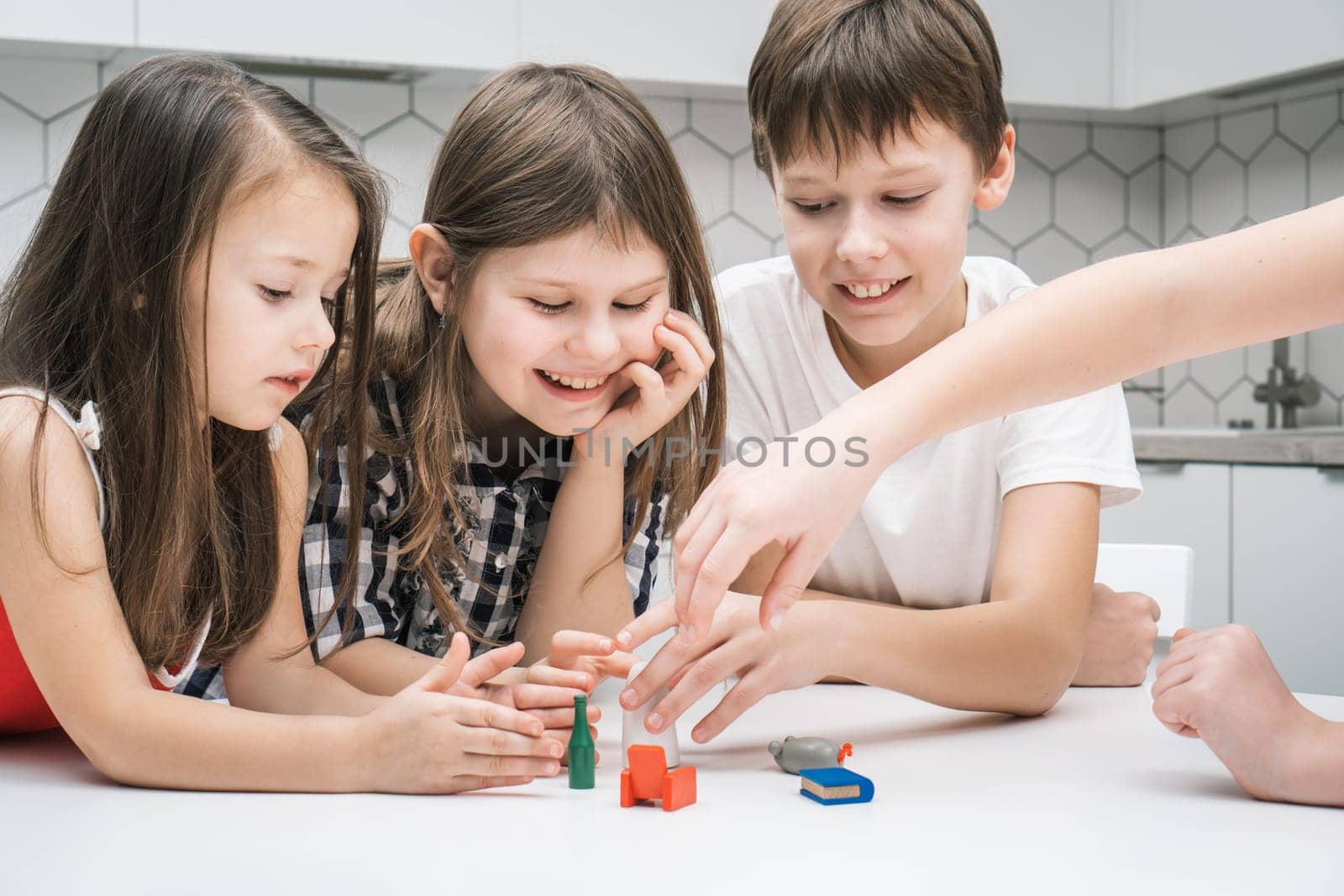 Happy little school kids play toy mini furniture on kitchen table. Portrait of group of friends holding small plastic playthings. Home leisure, funny playtime. Entertaining interesting board game.