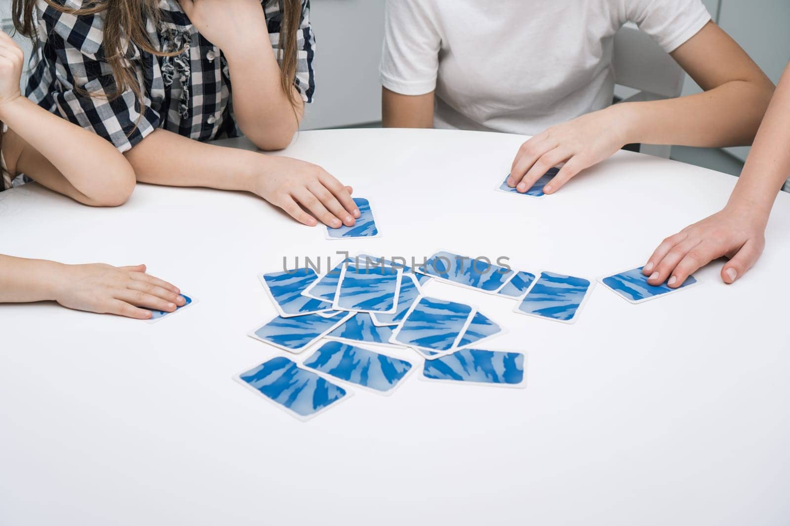Group of friends play card game sitting at white table. Children take with hands flipped paper blue cards from pile. Interesting entertaining board game for kids. Home leisure, funny playtime.