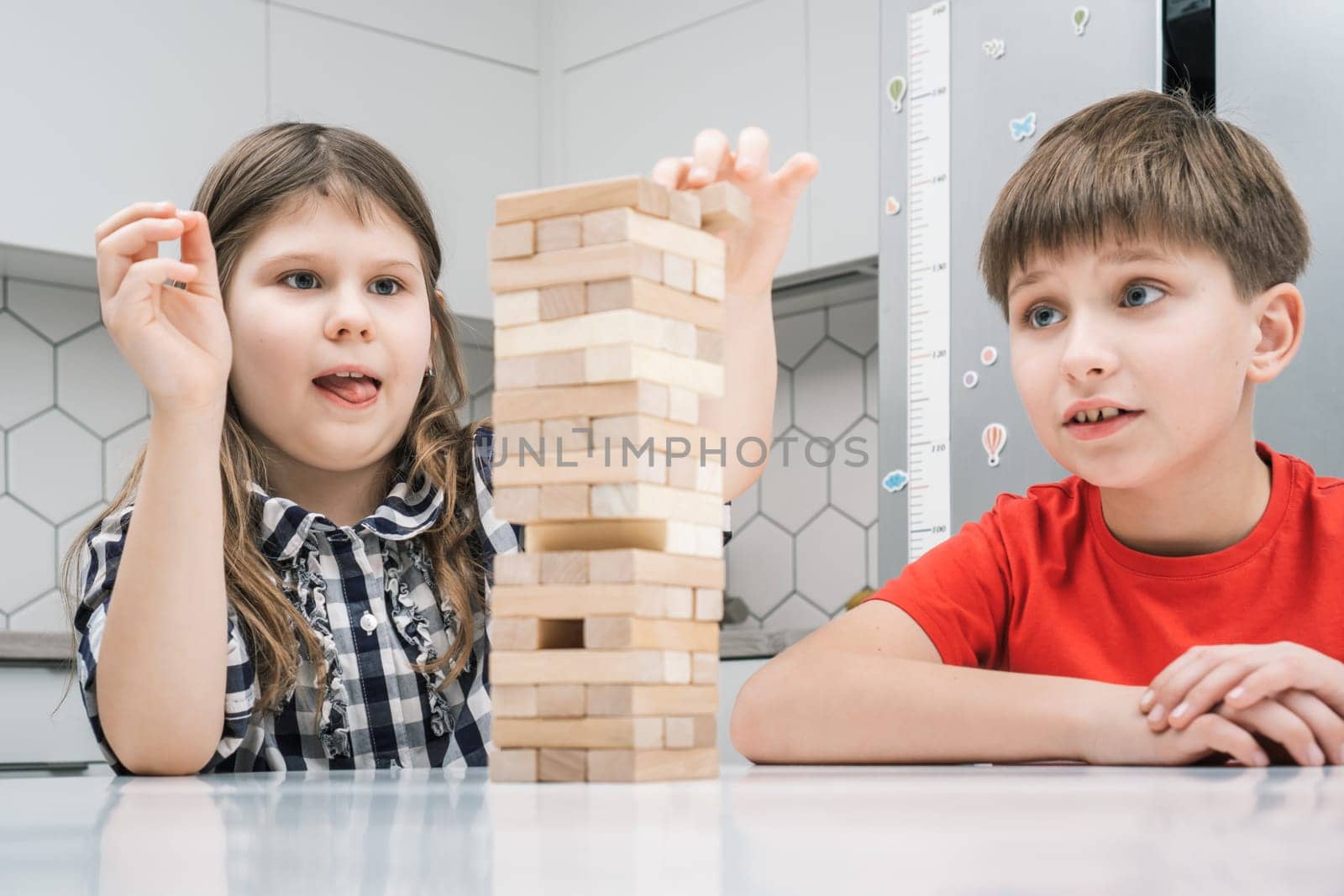 Cute school kids play jenga sitting at kitchen table. Boy and girl with concentrated facial expression build tower from wooden blocks. Home leisure, playtime. Entertaining interesting board game.