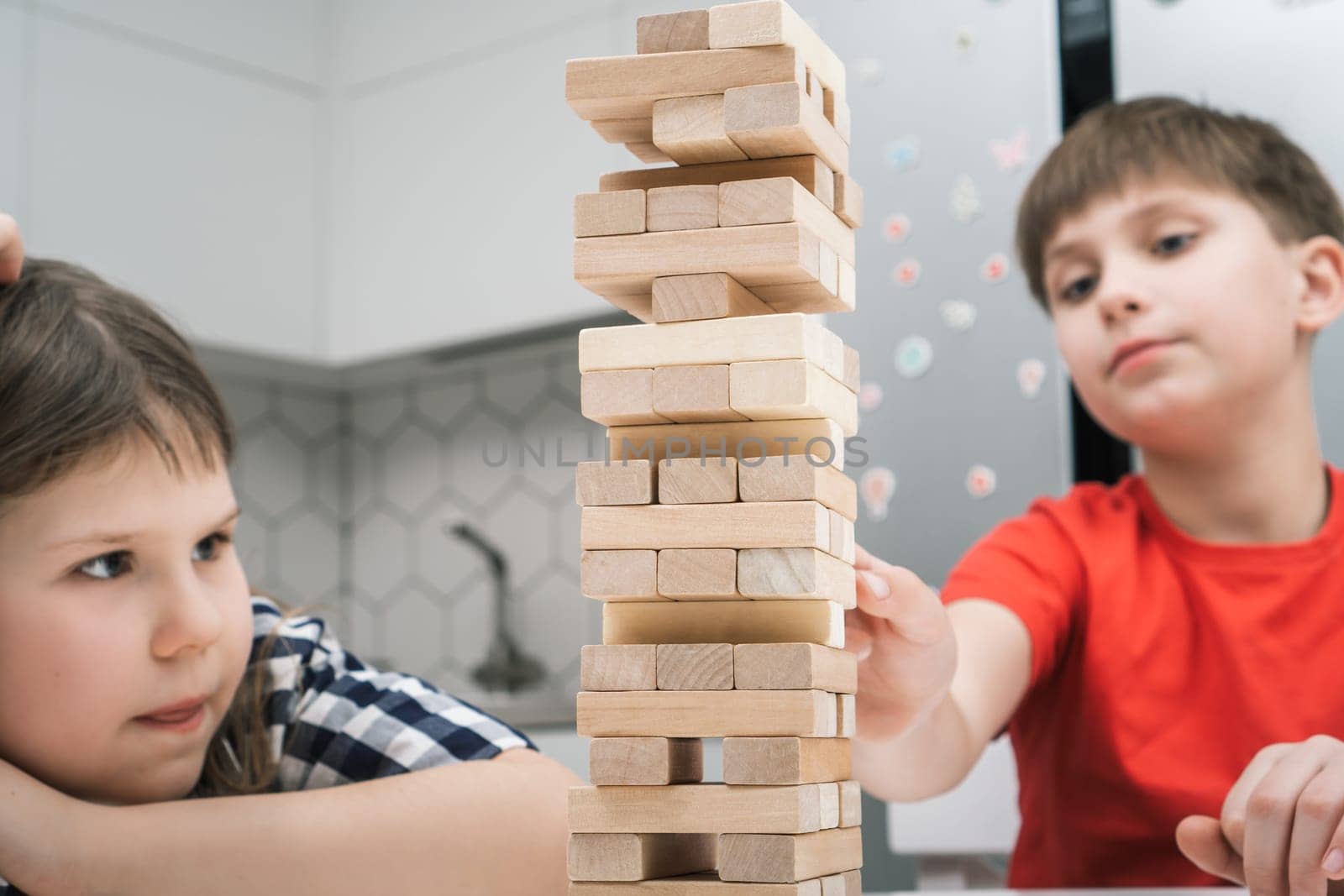 School kids play tower sitting at kitchen table closeup. Boy and girl build tower from little wooden blocks keeping balance. Friends relax at home, playtime. Entertaining interesting board game.