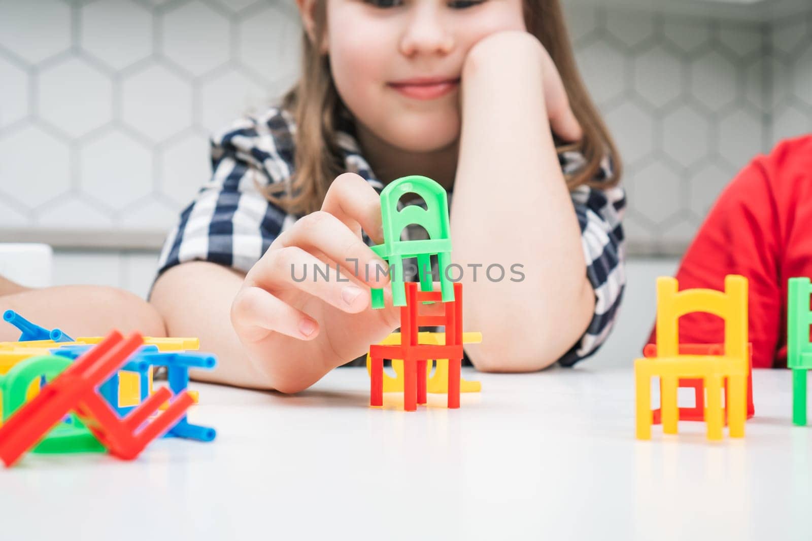 Little school girl play toy colorful chairs sitting at table closeup. Portrait of child holding small plastic furniture. Home leisure, playtime. Entertaining interesting board game.