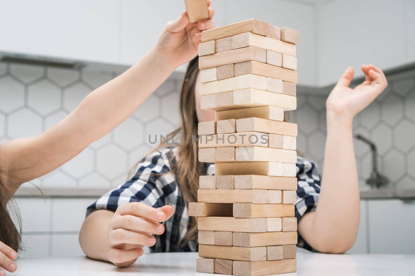 School kids play jenga sitting at kitchen table closeup. Hand of girl put little wooden block on top of tower keeping balance. Friends relax at home, playtime. Entertaining interesting board game.