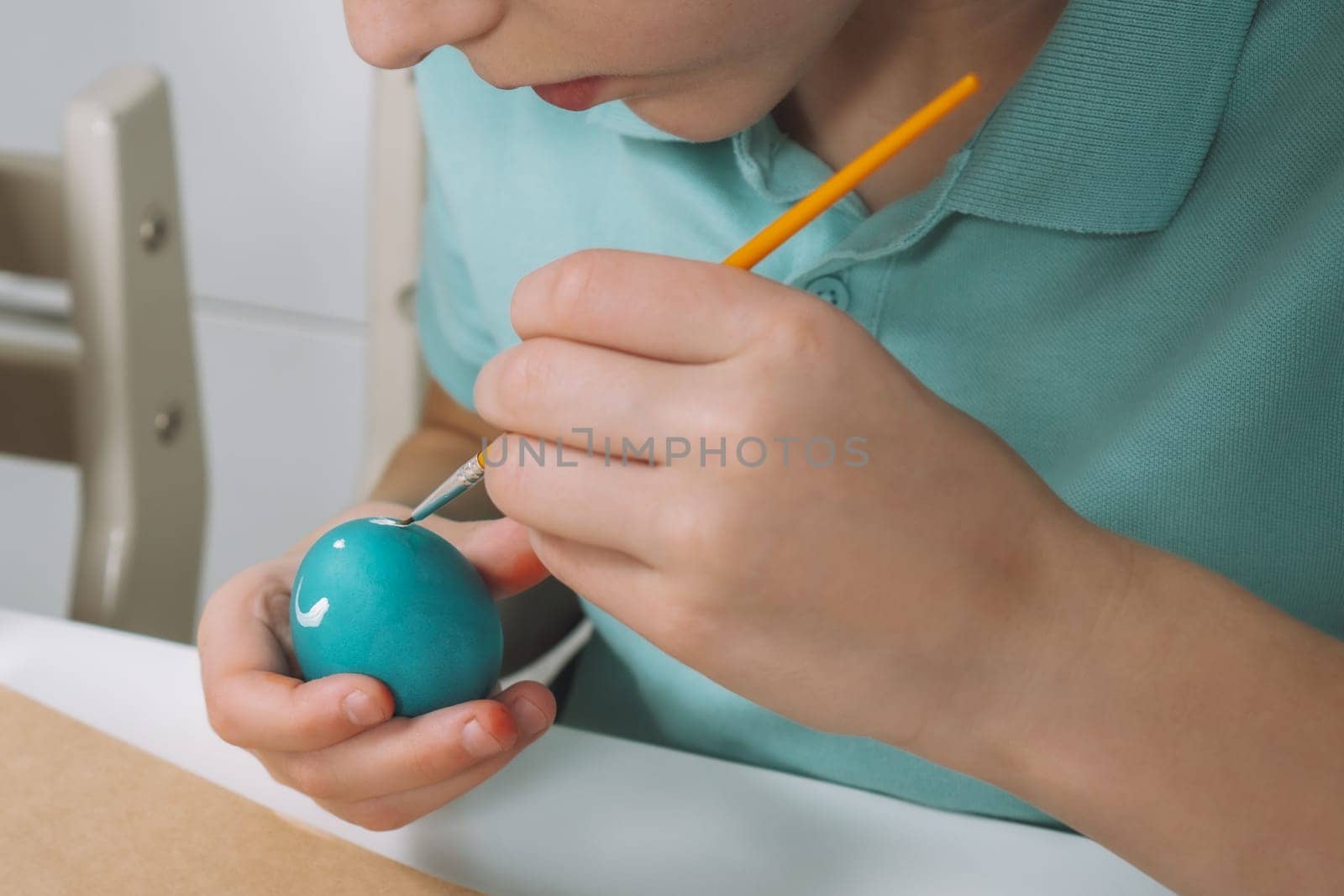 Cropped portrait of boy hand, painting multi coloring Easter hens eggs on brown paper plate table with paintbrush. Making art design white patterns on blue, red animal eggs. Holiday event. Close up