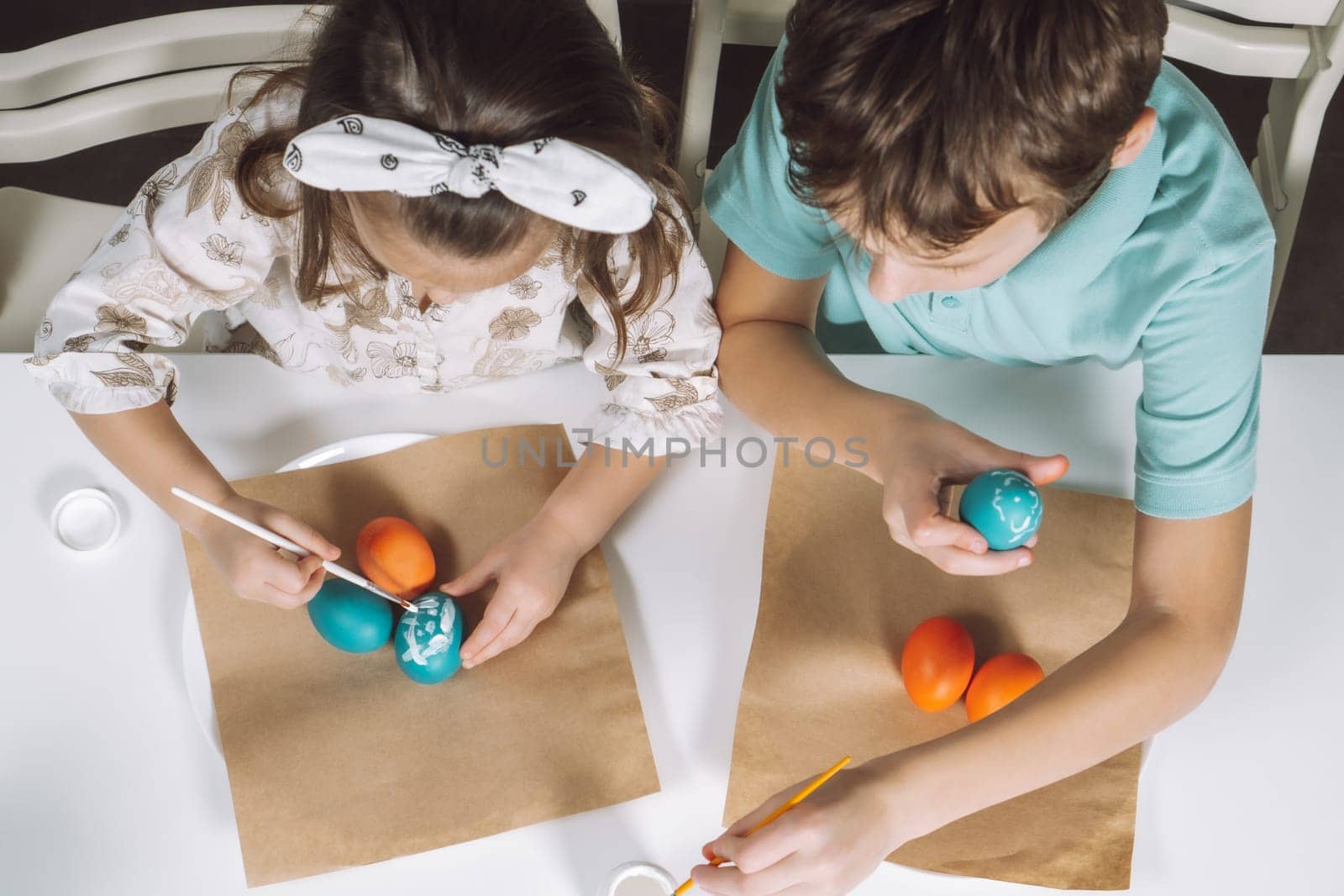 Portrait of two children, little girl and boy siblings, painting multi coloring Easter hens eggs in kitchen, with paintbrushes, sitting chair, paper table. Making art design white patterns. Top view.