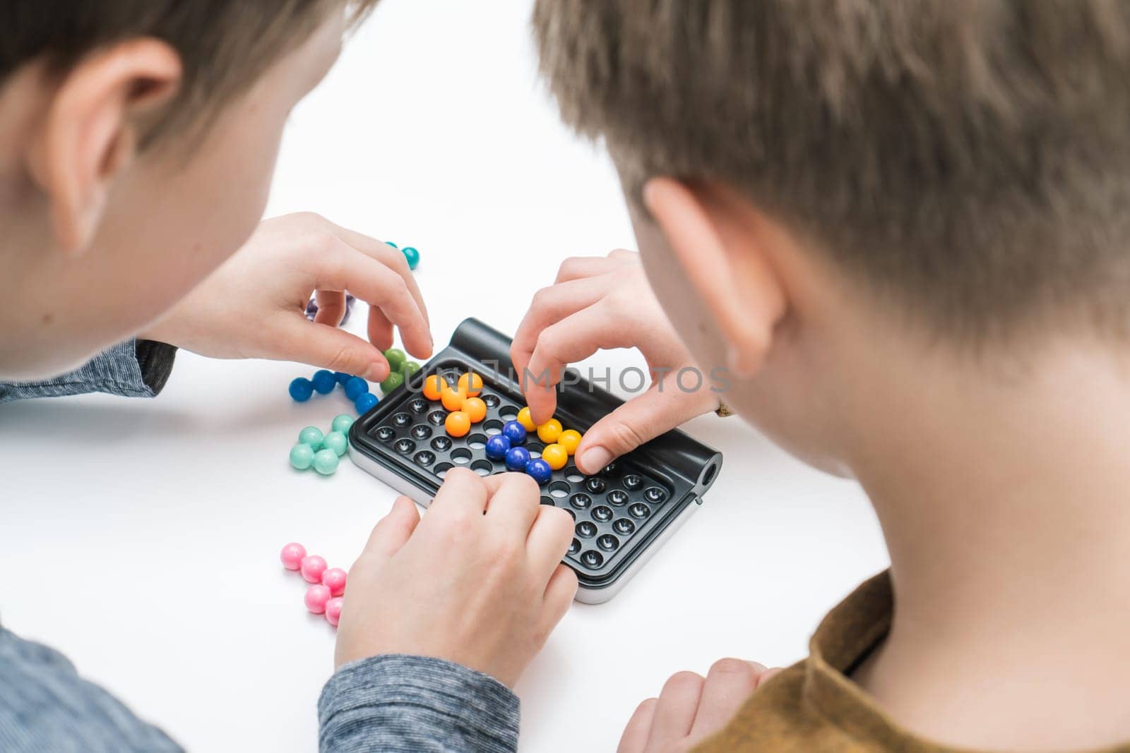 Boys play table tetris on table, from nape view. Children collect colored details on playing board. Hands of players placing figures on board closeup. Educational interesting board game.