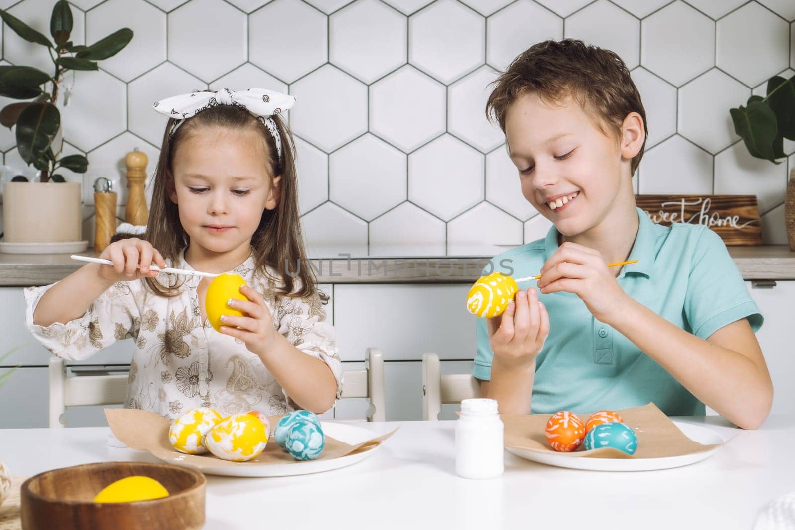 Portrait of concentrated joyful two children kids little girl and boy siblings, painting multi coloring Easter hens eggs in kitchen on paper table with paintbrushes. Making art design white patterns.
