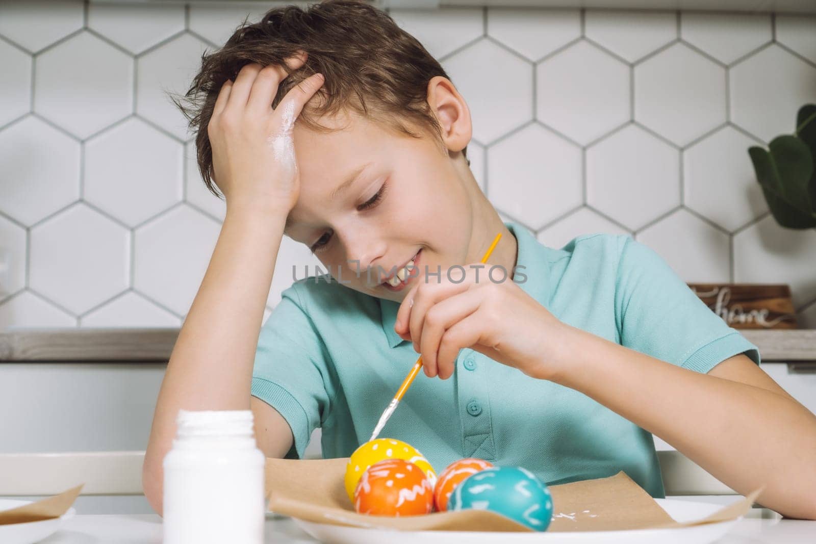 Portrait smiling joyful little boy, painting multi coloring Easter hens eggs paper table with paintbrush. Making art design white patterns. Sitting chair kitchen. Sweet home nameplate drawer
