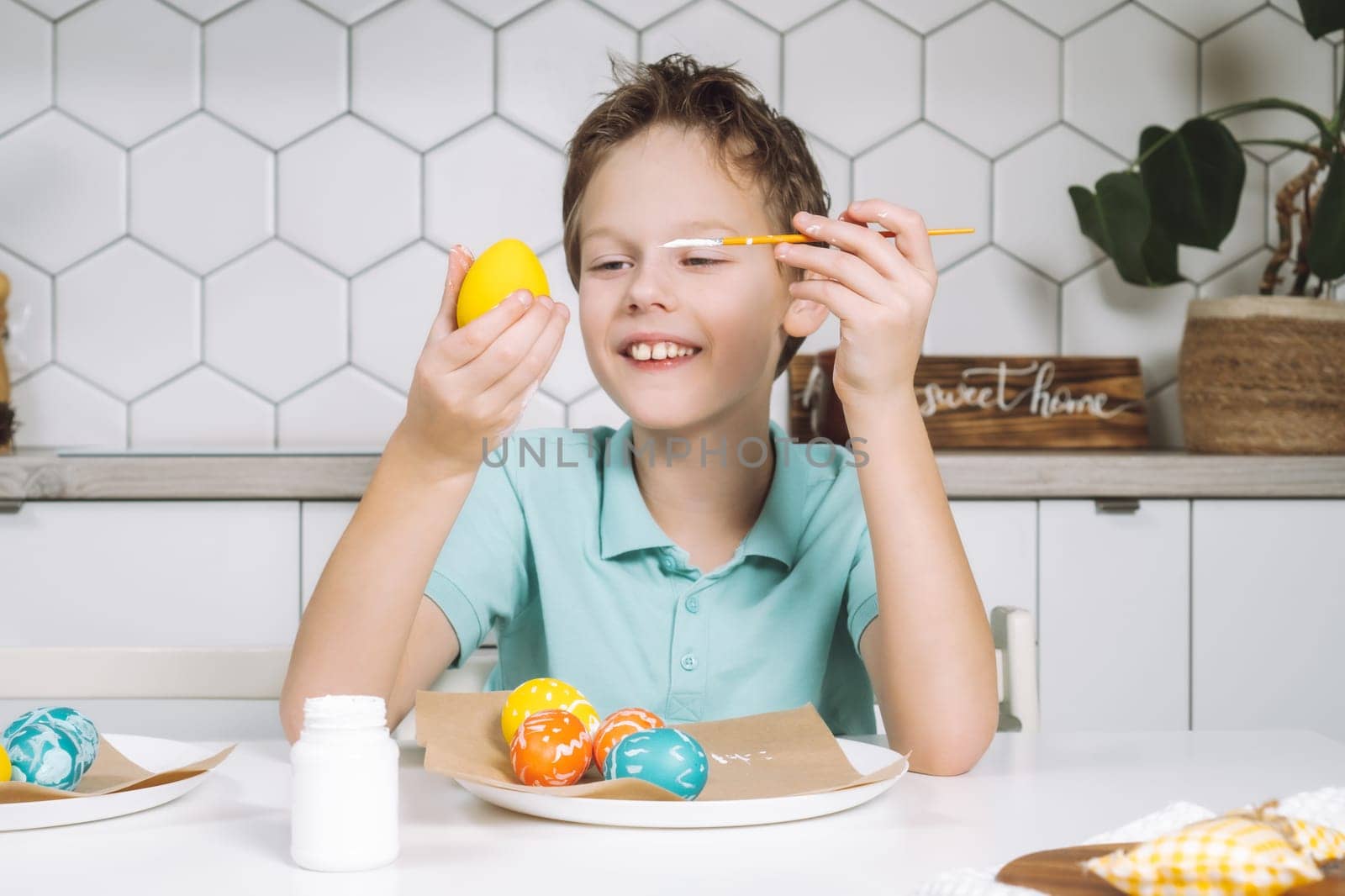 Portrait smiling joyful little boy, painting multi coloring Easter hens eggs paper table with paintbrush. Making art design white patterns. Sitting chair kitchen. Sweet home nameplate drawer