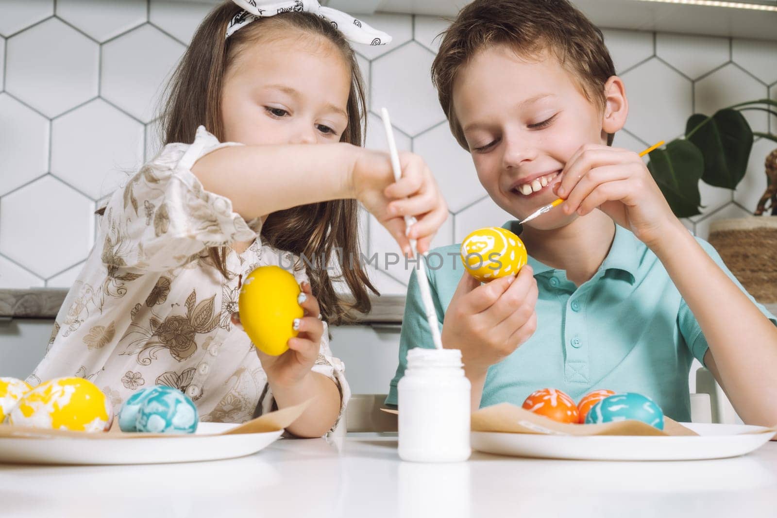 Portrait of concentrated joyful two children kids little girl and boy siblings, painting multi coloring Easter hens eggs in kitchen on paper table with paintbrushes. Making art design white patterns.