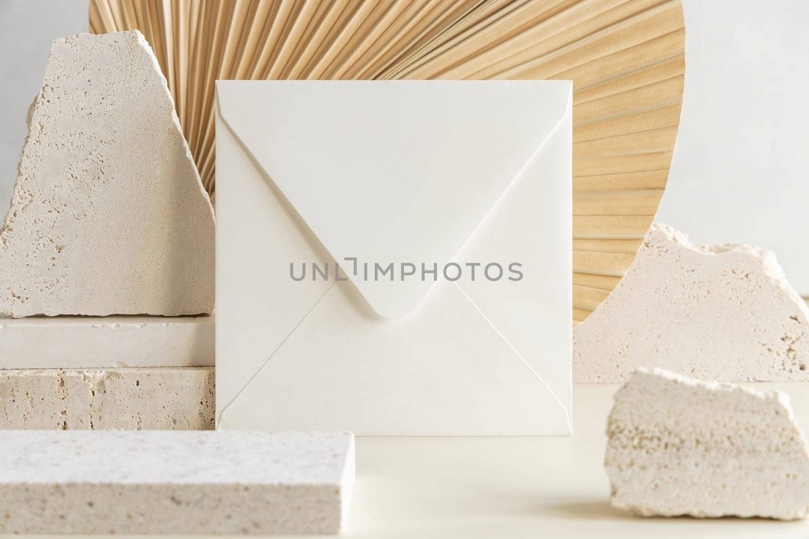 Blank square envelope near beige travertine stones against dry beige palm leaf close up, mockup. Boho scene with horizontal paper card. Bohemian greeting or wedding Invitation