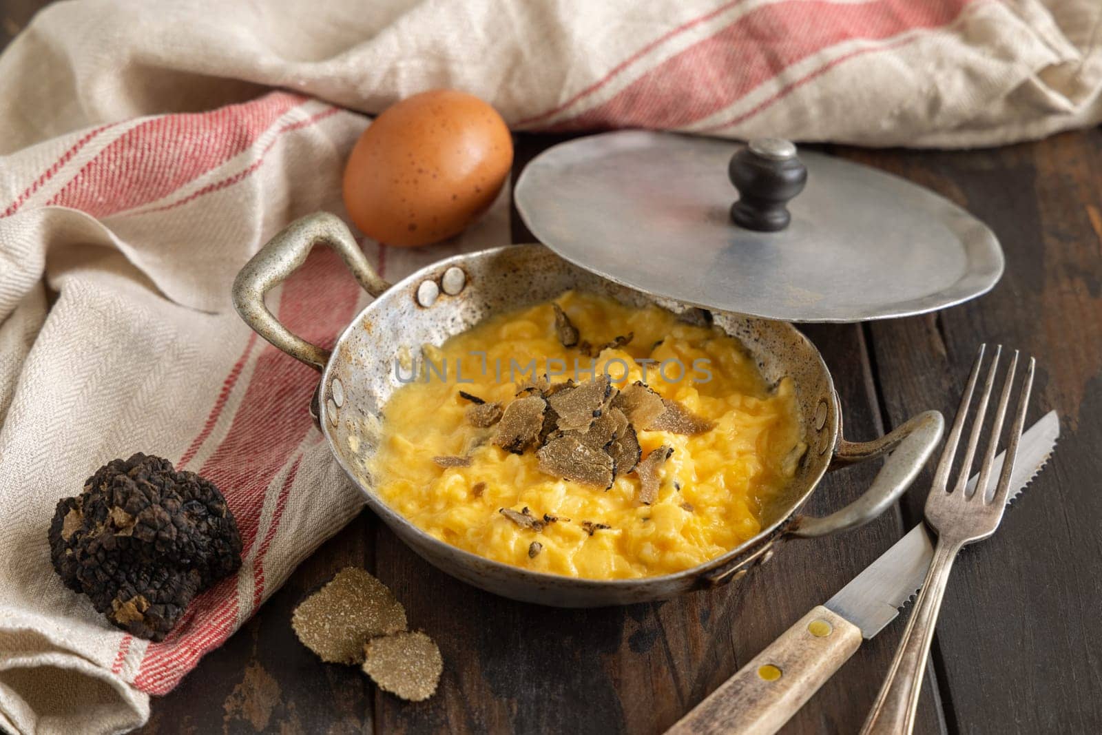 Scrambled eggs with sliced fresh black truffles from Italy served in a a frying pan near fork and knife on wood close up. Italian cousine, gourmet breakfast