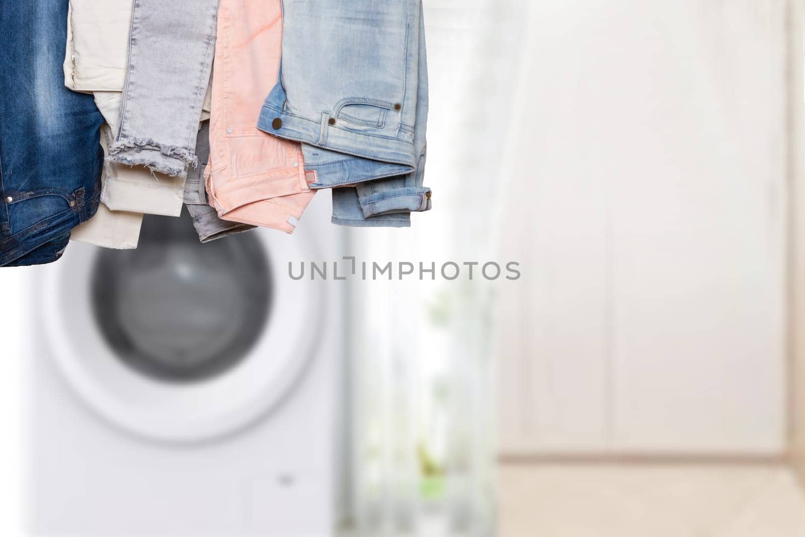 Hanging collection of female various denim pants or colorful trousers in front of abstract blurred bath background with copy space. For your product display montage.