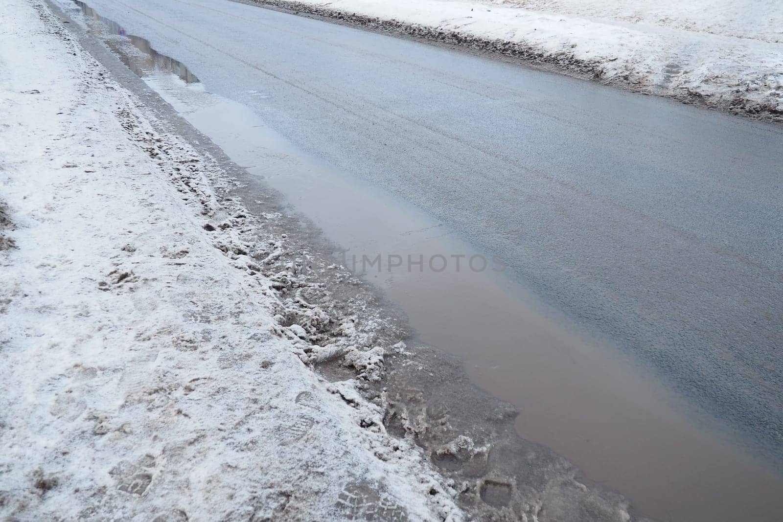 Ice on city roads. Wet asphalt with sand. Snow-covered roadsides. Weather forecast. Thaw in winter. Snow melting. The air temperature is near zero. Footprints of people. Difficult driving conditions