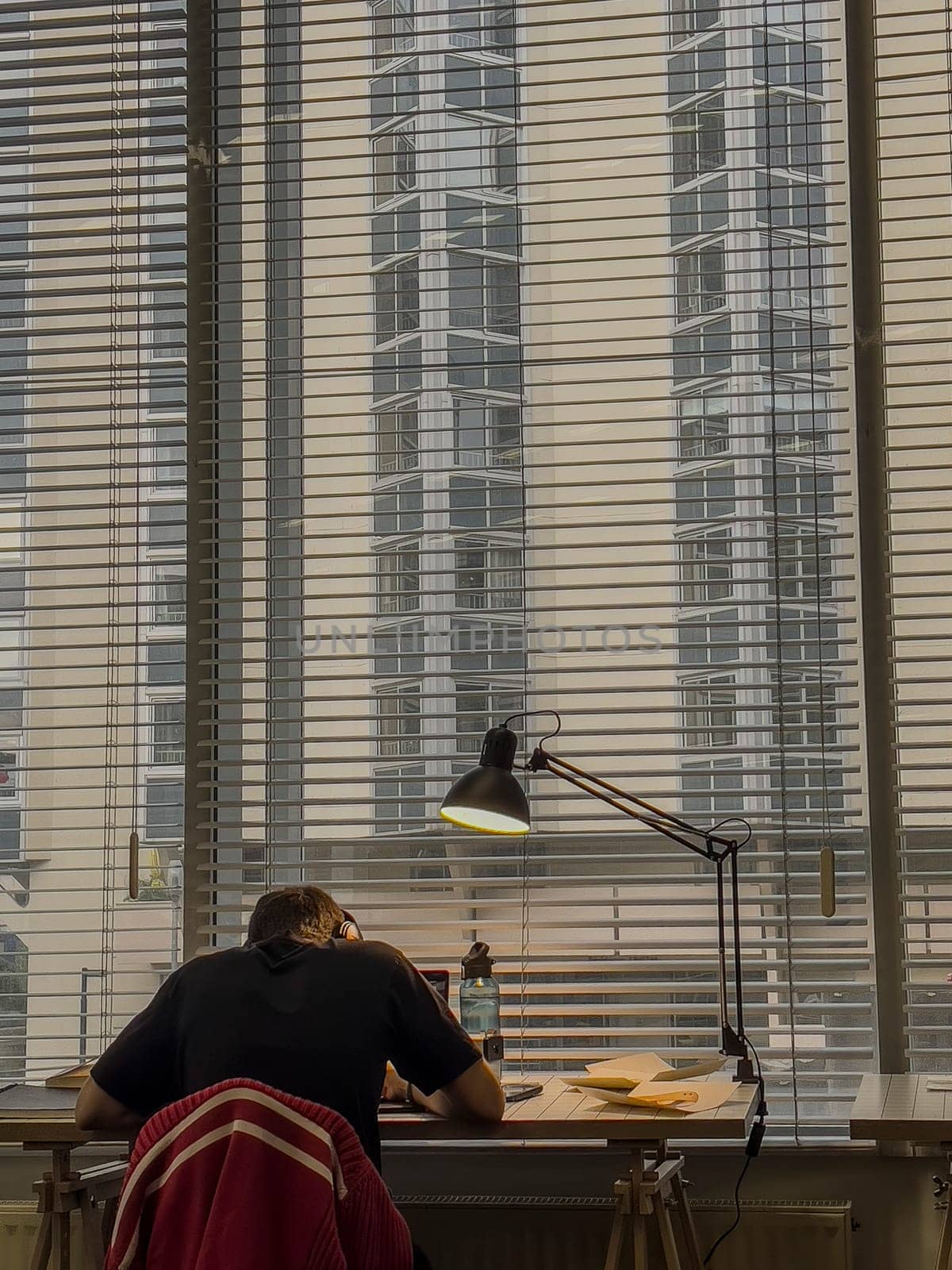 Inside a contemporary office, three students focus on their work at desks under bright daylight streaming through tall windows showcasing a city view.