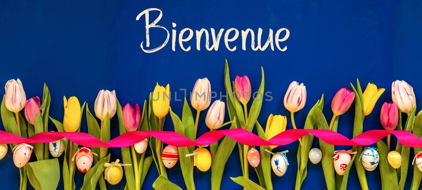 French Text Bienvenue Means Welcome. Banner Of White And Pink Tulip Spring Flowers With Ribbon And Easter Egg Decoration. Blue Wooden Background