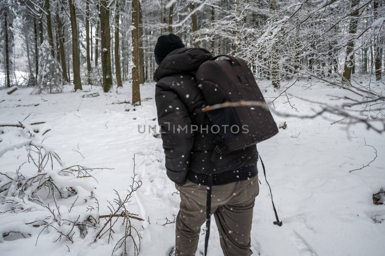 Man with hat and backpack is hiking trough the forest during winter with lots of snow, rear view