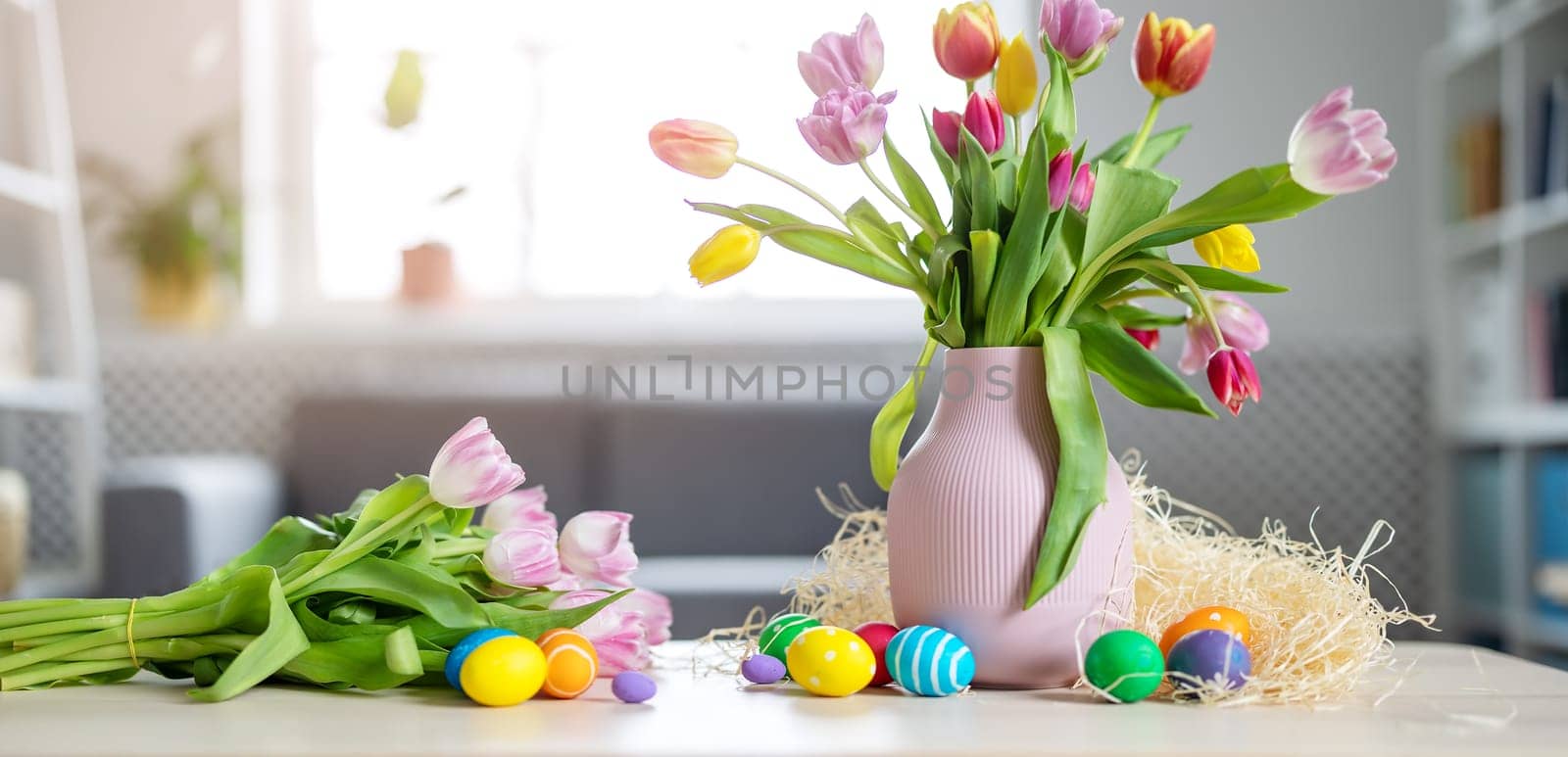 A bunch of tulips in a vase standing on the table and colourful eggs lying on it. Concept of the Easter.