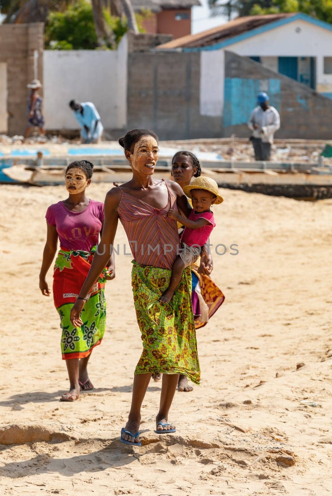 Morondava, Madagascar - November 3. 2022: Malagasy woman with her face painted against the sun and a child in her arms walks along the beach in Morondava.