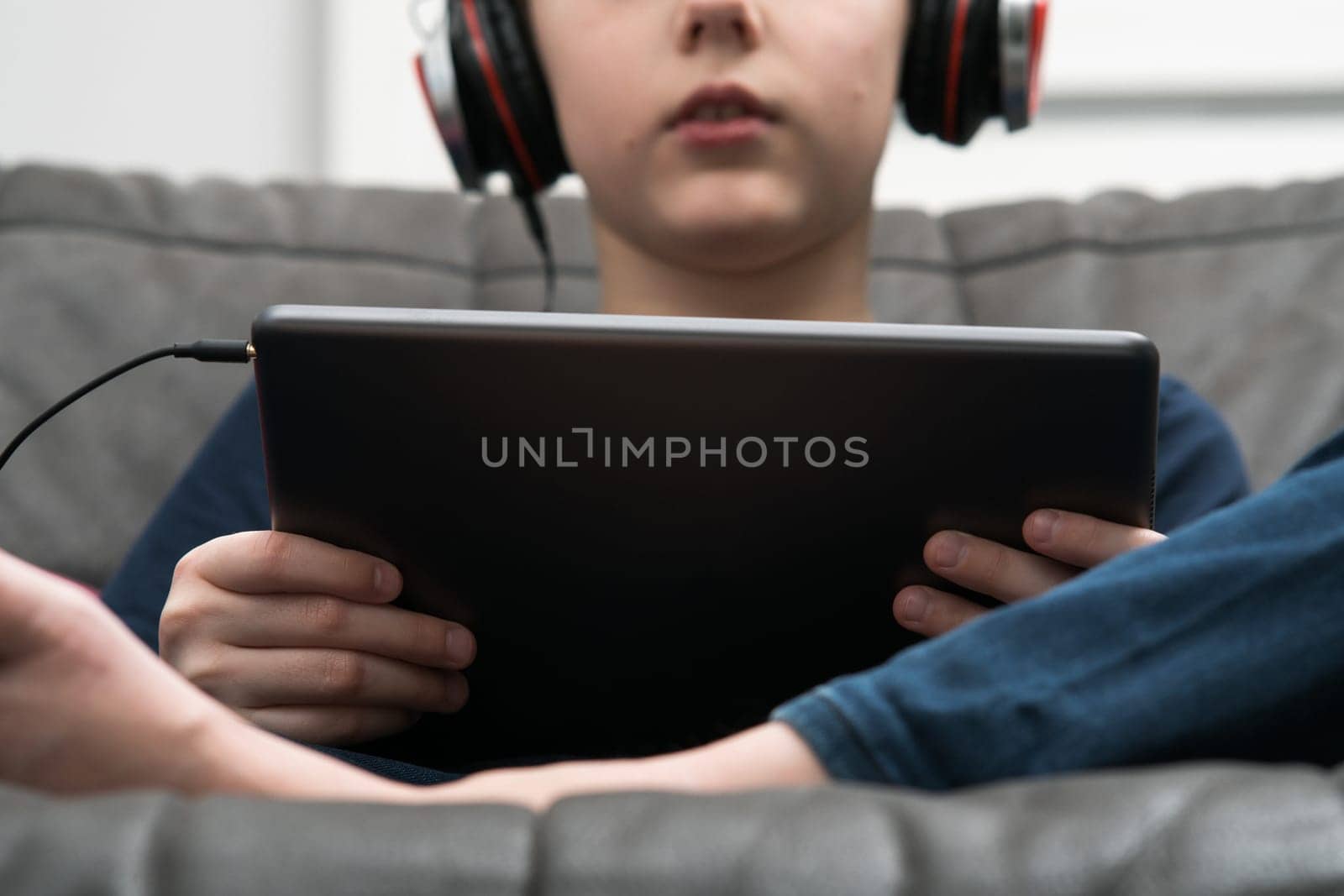 Cropped midsection of little boy in wired headphones connected with digital tablet in hands, sitting on sofa. Online courses, homeschooling and interactive lessons platform online. Music and movies