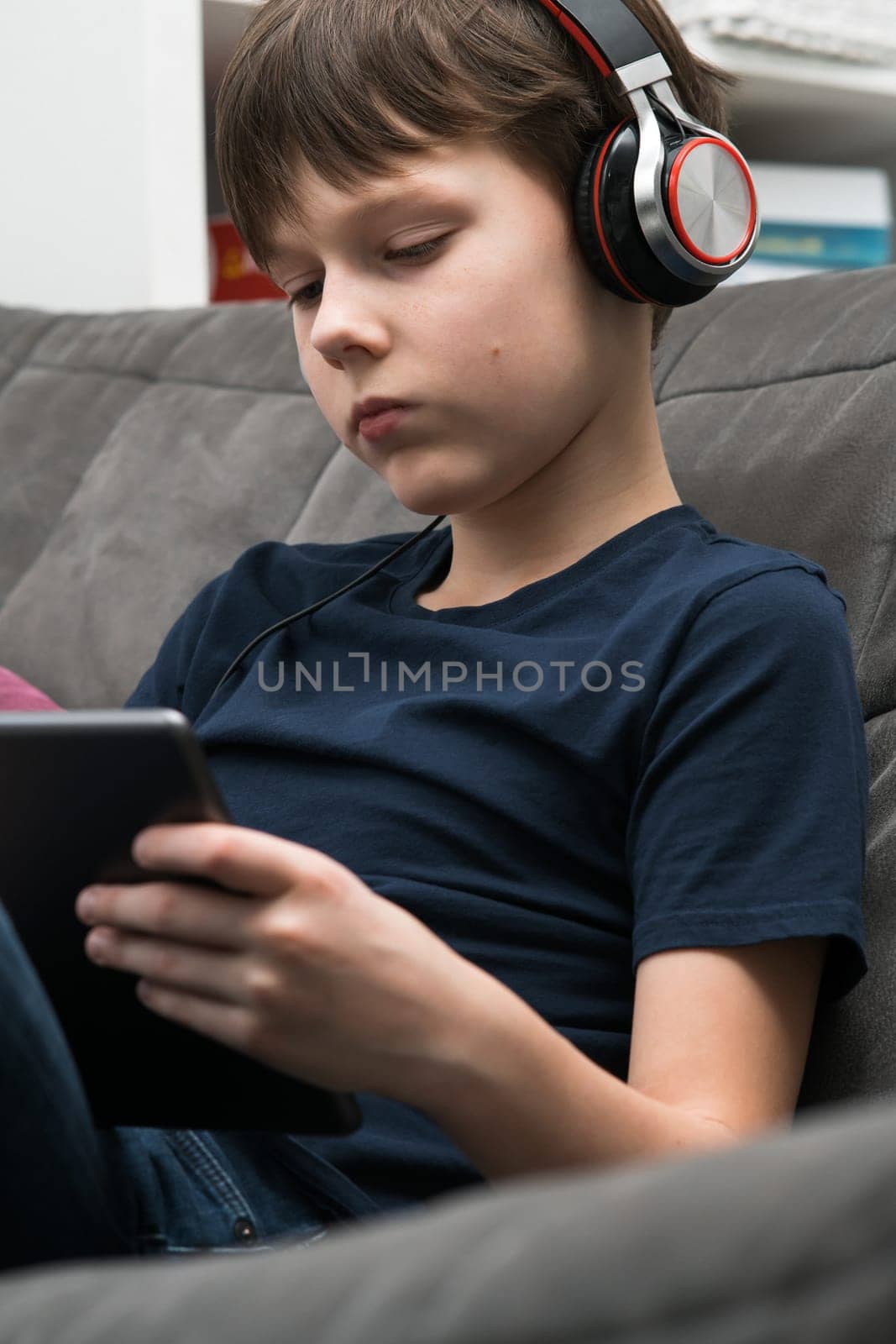 Vertical tranquil, concentrated little boy in headphones listen to music and watching movies with digital tablet, pad in hands, sitting on sofa. Portable Bluetooth device. Podcasts in your earphones.
