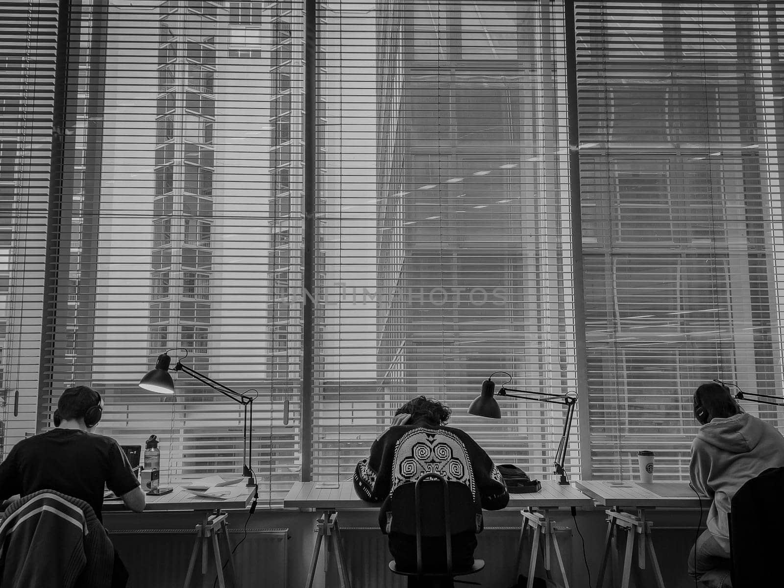 Inside a contemporary office, three students focus on their work at desks under bright daylight streaming through tall windows showcasing a city view.