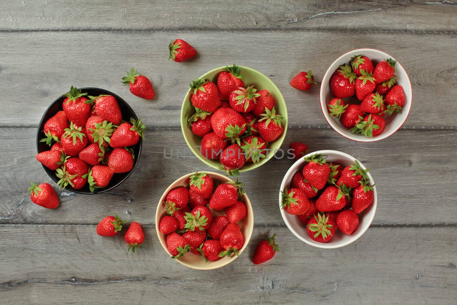 Tabletop view, small bowls with strawberries, some of them spilled on gray wood desk.