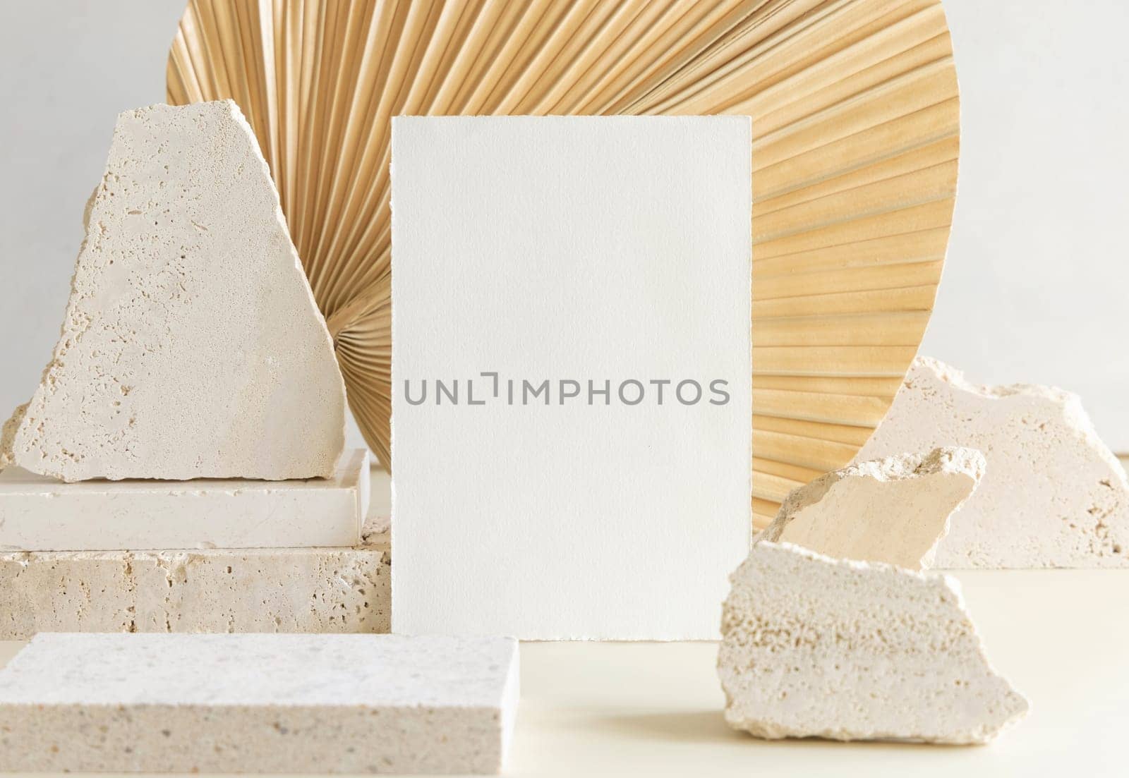 Blank card near beige travertine stones against dry beige palm leaf close up, mockup. Boho scene with vertical paper card. Bohemian or close to nature greeting or wedding Invitation. Minimal composition.