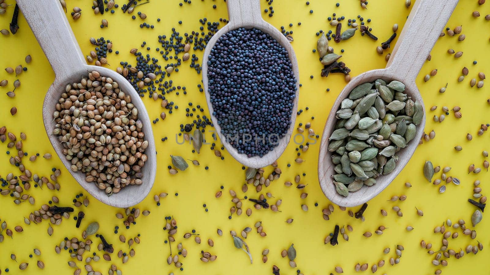 Black mustard seeds, coriander and cardamom in wooden spoon on yellow background from above