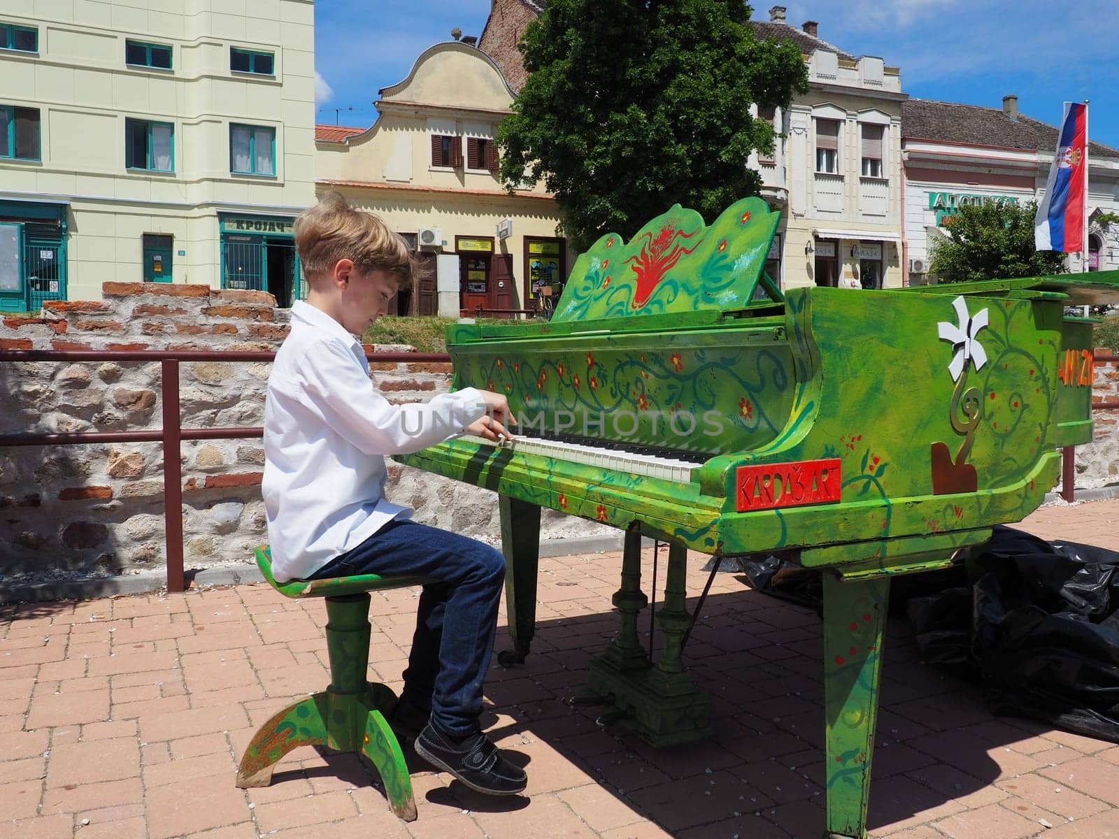 Sremska Mitrovica, Serbia, 05 June 2021. A boy in a white shirt plays the piano in the street. The child is 9 years old Caucasian. Piano painted green. Performance of a piece of music. Street concert.
