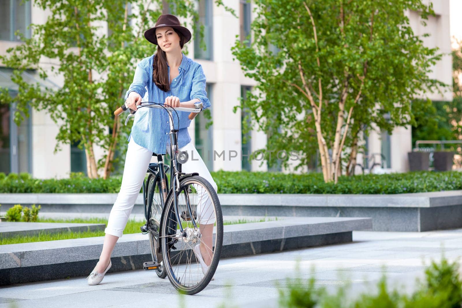 Pretty smiling girl in hat and blue dress riding a bicycle at green street of town