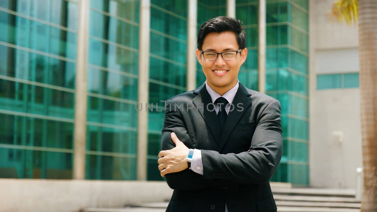 Portrait of happy and confident young asian businessman. The man stands in a street against office buildings and crosses arms looking at camera