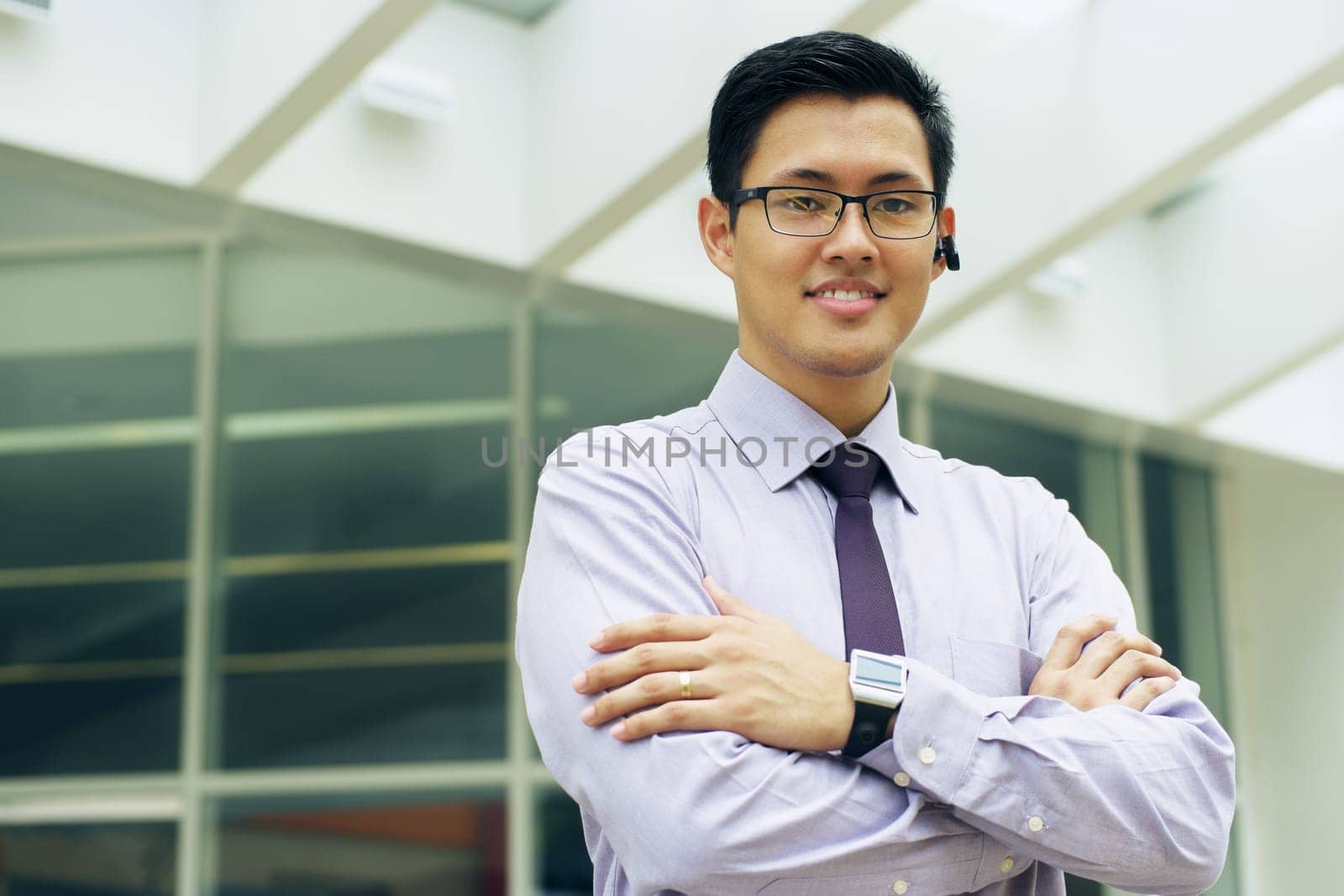 Portrait of confident asian businessman. The man stands in a street against office buildings and crosses arms looking at camera with bluetooth headset and smartwatch
