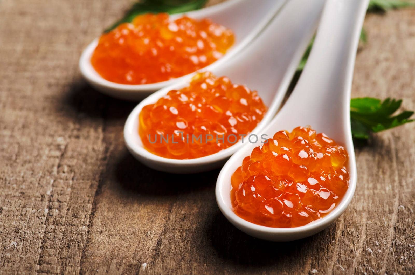 Closeup of luxury red caviar in white spoon with parsley on wooden table background. Original russian seafood cuisine.