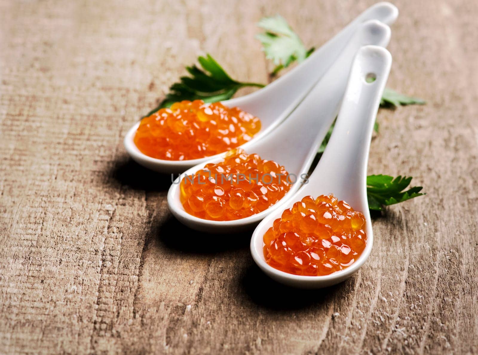 Closeup of luxury red caviar in white spoon with parsley on wooden table background. Original russian seafood cuisine.