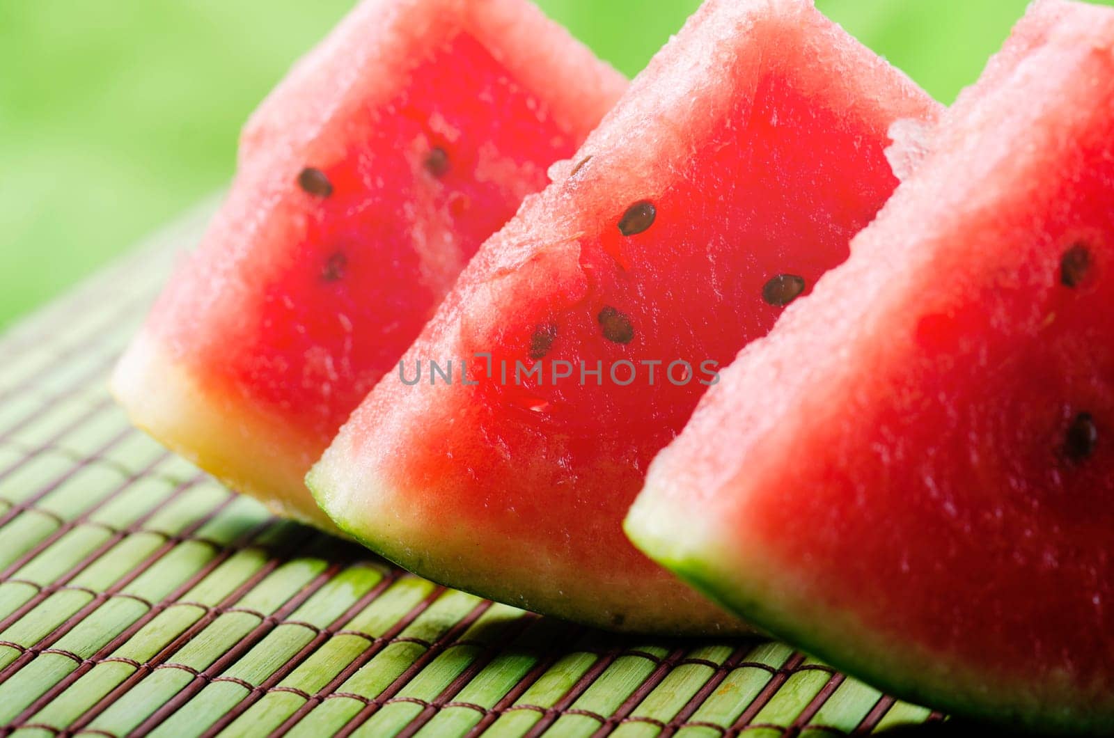 Closeup of sweet red watermelon slices on green table background in garden with sunlight. Summertime refreshing sweet food. Colorful fruit still life..