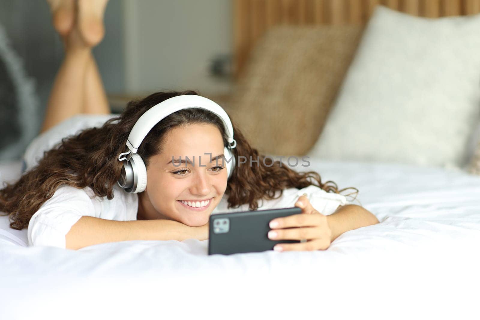 Happy woman wearing headphones watching videos on smart phone lying on a bed in a bedroom