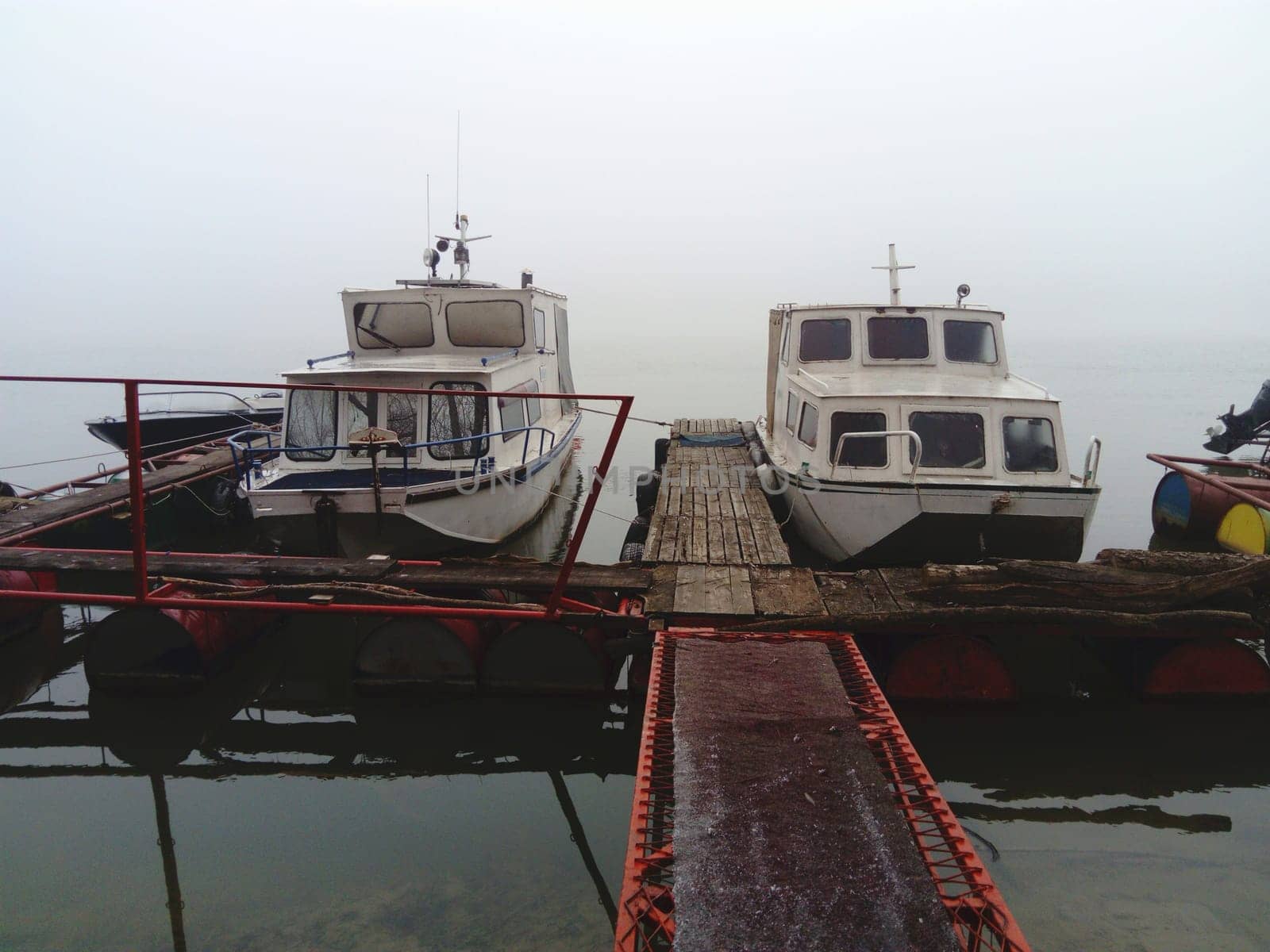 Belgrade, Serbia, January 14, 2020 Two white boats at a wooden pier, left for the winter. Foggy weather in southern Europe. January cloudy day. Reflection of water transport in a river river.