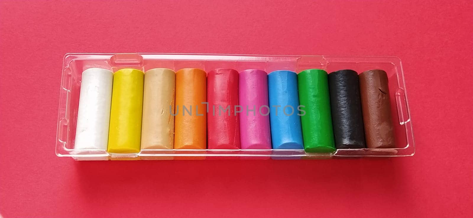 Multi-colored new plasticine in plastic packaging on a red background. Plasticine plates of red, pink, green, white, yellow, brown, blue, black. Set for children's creativity.