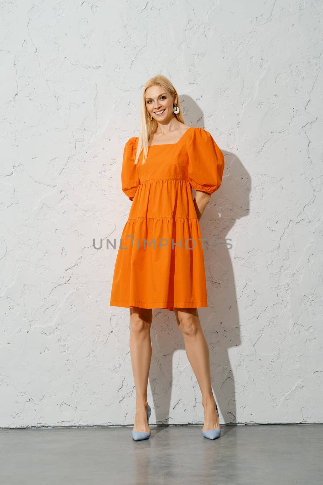 A smiling woman with her hands behind back in a bright orange spring dress stands indoors, posing for the camera