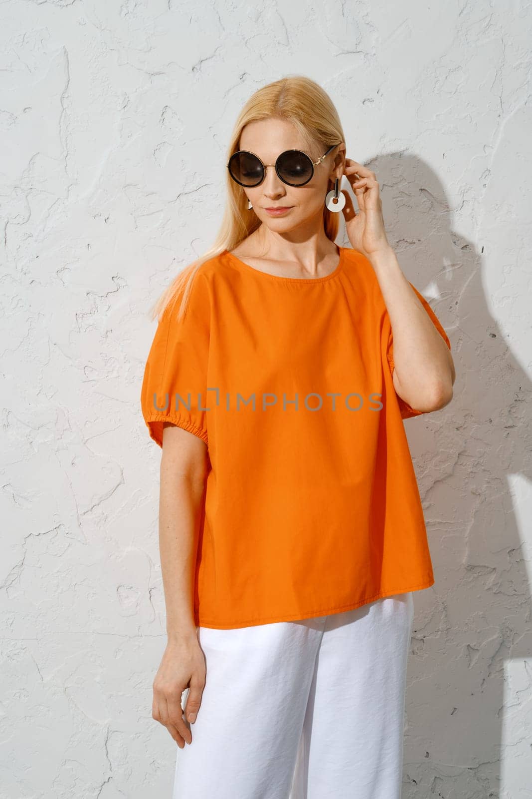 A fashionable woman stands in three-quarter length, wearing an orange top with short sleeves and sunglasses