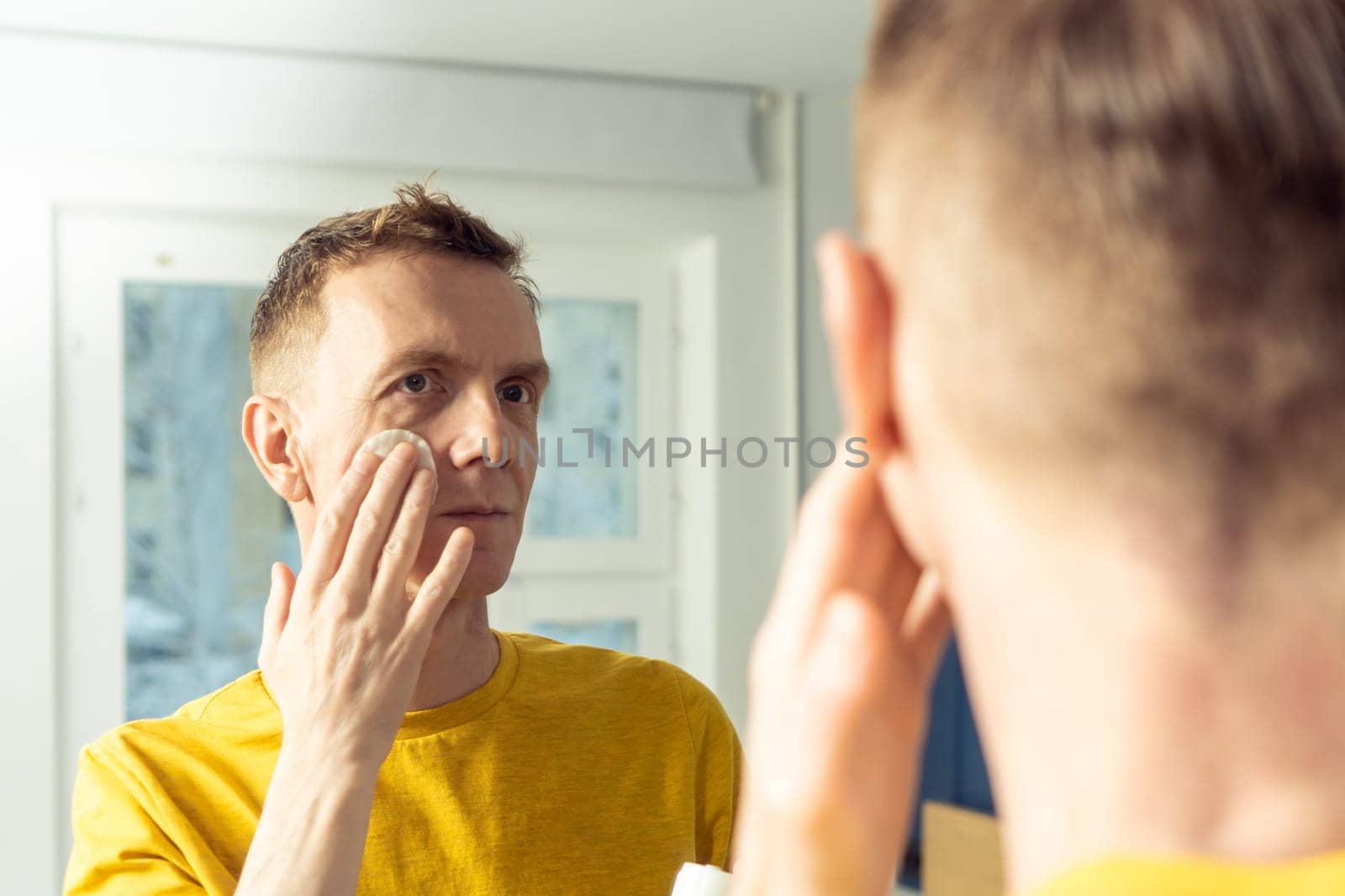 Middle aged man apply face tonic with cotton pad, then massage with hands. Male portrait in mirror. Skincare morning routine, careful beauty treatment, home lifestyle. Face cleansing, washing.