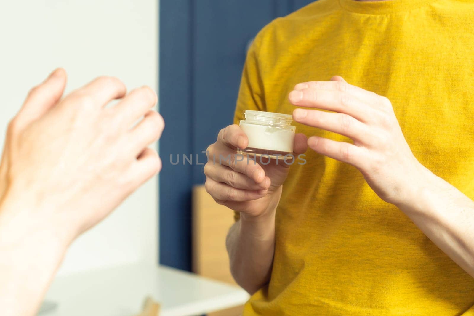 Hands of middle aged man holding jar of moisturizing cream. Crop male portrait in room mirror. Skincare morning routine, careful beauty treatment, home lifestyle. Facial care concept.