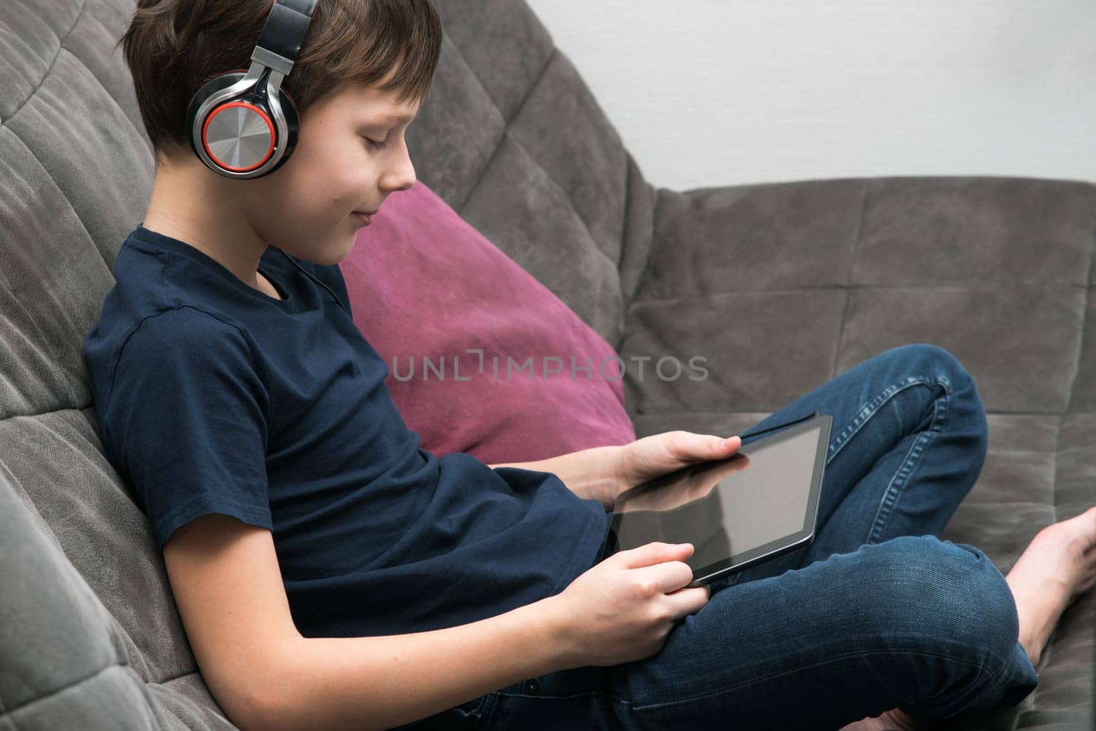 Portrait of happy little boy with digital tablet computer resting on sofa, side view. Child in headphones play videogame on gadget or watch video. Online entertainment, home leisure, gamer.