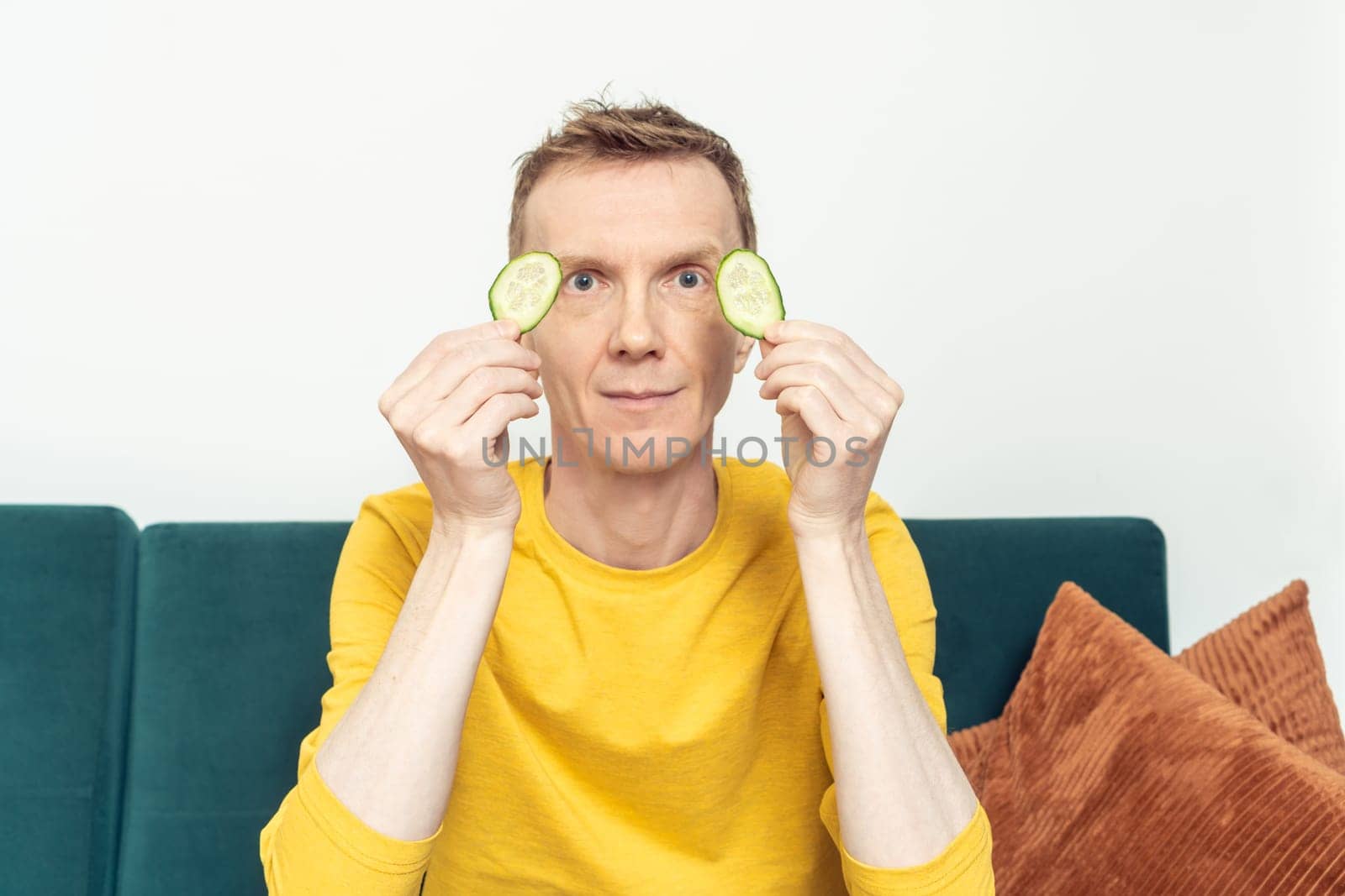 Happy adult man sitting on sofa hold in hands eye patches from fresh cucumber slices in living room. Mature person do moisturizing mask for dark circles under eyes. Home lifestyle, beauty treatment.