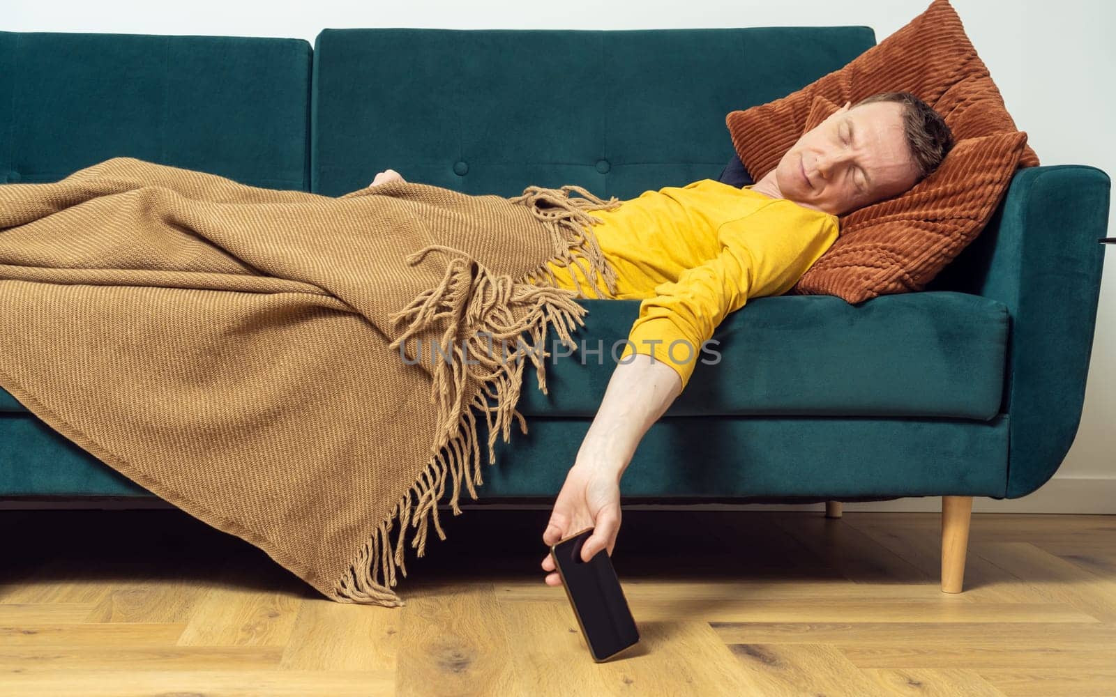 Tired adult man sleep on sofa with smartphone in hanging hand in living room. Overworking person reclining on pillow under blanket, side view. Home relaxation, rest break, leisure, lounge.