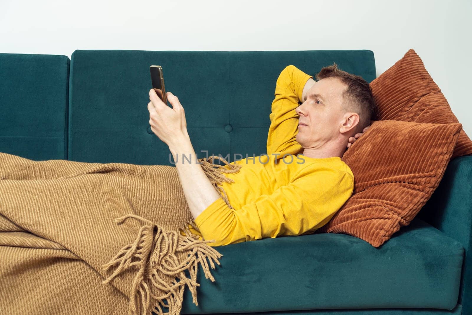Adult man lie on sofa and chatting on smartphone in living room, side view. Person reclining on pillow under blanket and lounging. Look at screen of phone and read. Home relaxation, lazy activity.