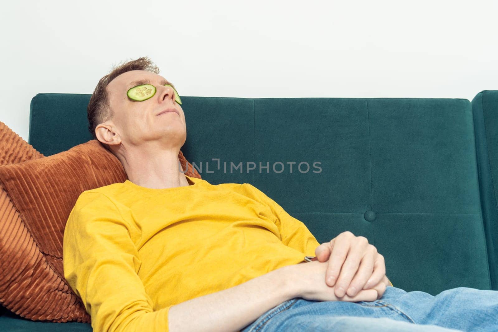 Adult man lying on sofa with eye patches from fresh cucumber slices in living room. Middle aged person reclining on pillow with mask for dark circles under eyes. Home relaxation, beauty treatment.