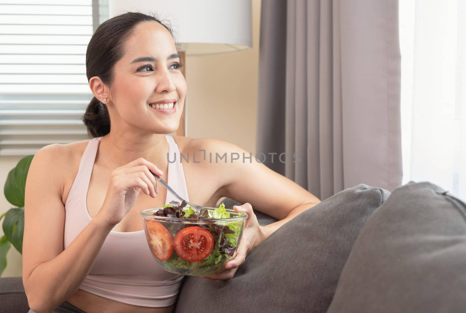 Young beauty woman wearing a sportive outfit and enjoy heathy meal with vegetable salad on sofa after yoga execise at home with happiness. Healthy lifestyle and exercise concept